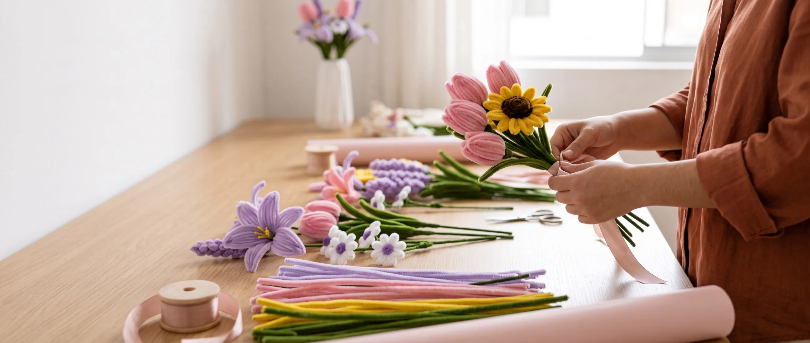 Unas manos atan con cinta un ramo pequeño de tulipanes y un girasol de chenille sobre una mesa de trabajo llena de flores y tallos de colores