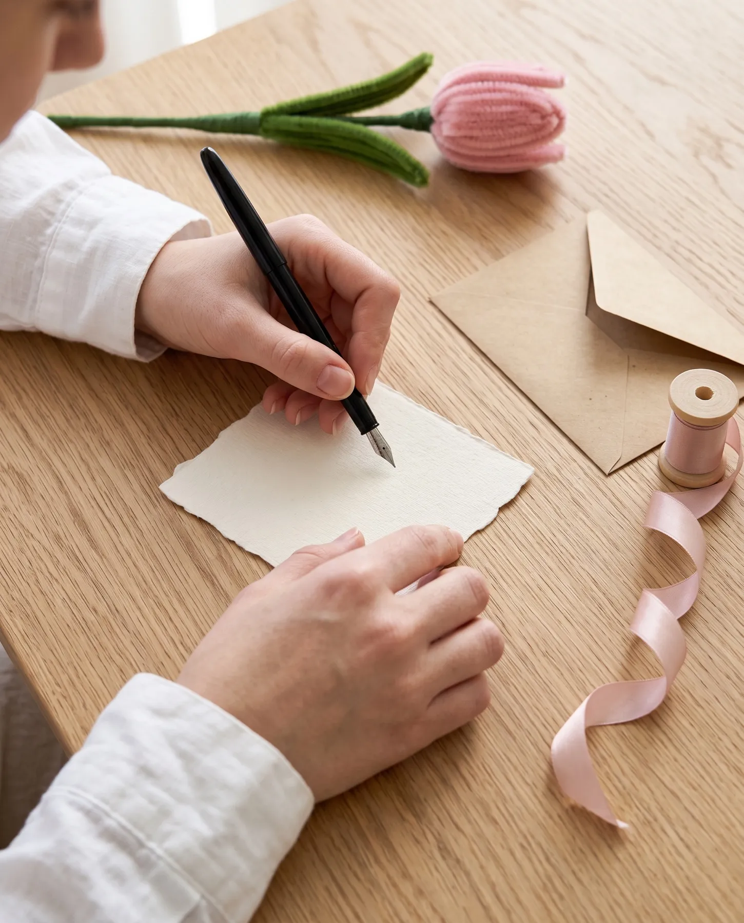 Una mano escribe una tarjeta de dedicatoria junto a un sobre kraft y una cinta rosada